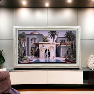 May include: A large rectangular television screen framed in an ornate white decorative border, displaying a vibrant digital image of a desert-style villa with palm trees, a swimming pool, and white birds, mounted above a sleek white media console.