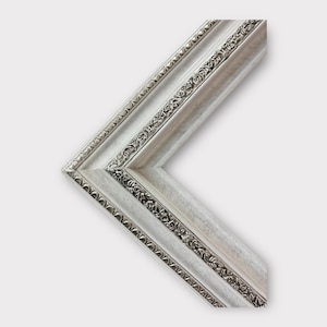 May include: A corner section of a silver picture frame. The frame has a detailed floral pattern along the inner and outer edges. The frame's color is a mix of silver and white.
