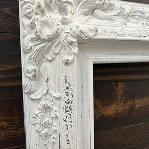 May include: Ornate white picture frame with distressed finish. The frame features detailed floral and scrollwork carvings, with hints of gold peeking through the white paint. The frame is rectangular and appears to be made of wood.