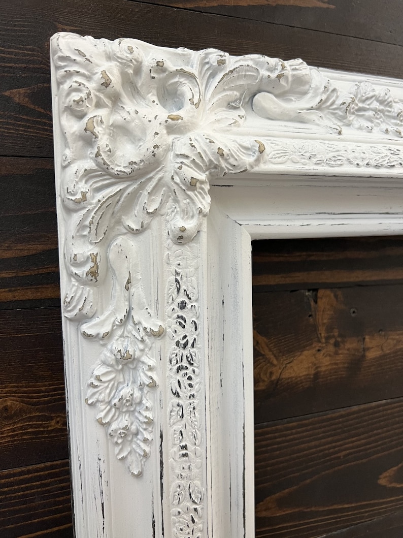May include: Ornate white picture frame with distressed finish. The frame features detailed floral and scrollwork carvings, with hints of gold peeking through the white paint. The frame is rectangular and appears to be made of wood.
