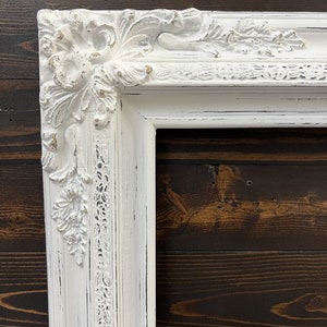 May include: A white, ornate picture frame with a distressed finish. The frame features detailed floral and scrollwork carvings at the corners and along the edges. The frame's design is classic and elegant, with a vintage aesthetic.
