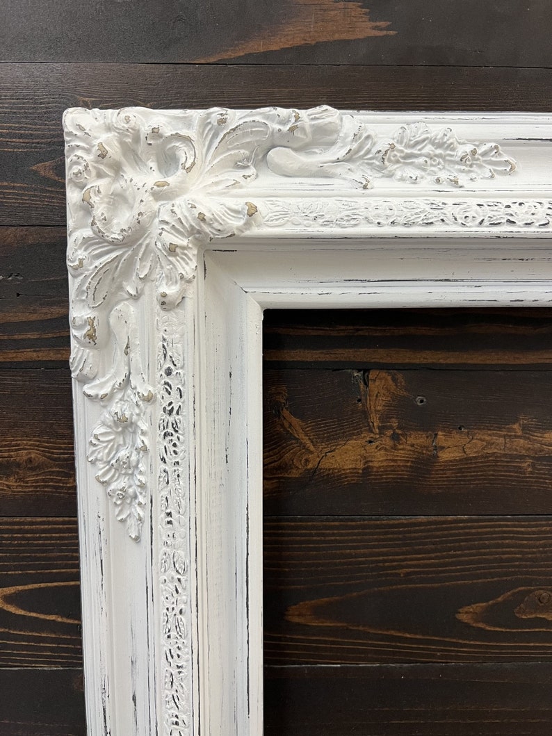 May include: A white, ornate picture frame with a distressed finish. The frame features detailed floral and scrollwork carvings at the corners and along the edges. The frame's design is classic and elegant, with a vintage aesthetic.