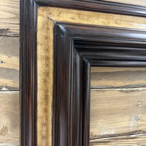 May include: Close-up of a rectangular picture frame with a dark brown exterior and a gold-colored interior border. The frame has a detailed, ornate design with vertical grooves and a glossy finish. The background is a wooden surface.