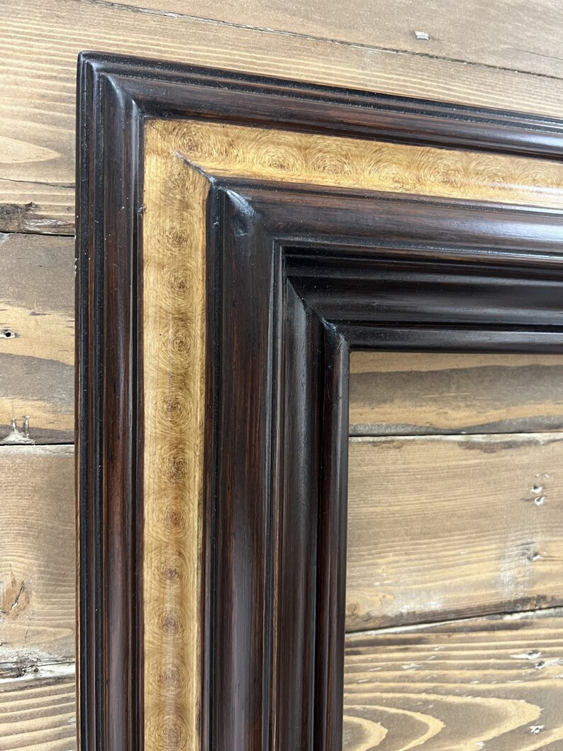 May include: Close-up of a rectangular picture frame with a dark brown exterior and a gold-colored interior border. The frame has a detailed, ornate design with vertical grooves and a glossy finish. The background is a wooden surface.