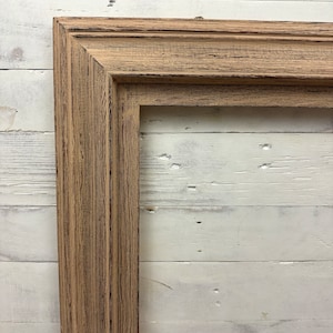 May include: A rectangular picture frame featuring a light brown wood finish with a visible grain texture and dark distressed accents. The frame has a multi-tiered stepped profile with clean mitered corners, showcasing a natural weathered appearance.