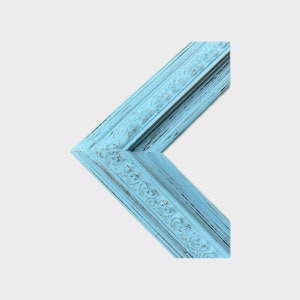 May include: A corner section of a picture frame featuring a light blue finish with a distressed, weathered texture. The frame has an ornate, raised floral relief pattern running along the center, showing dark wood undertones through the paint.