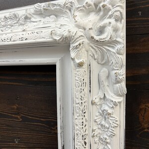 May include: Close-up of a white, ornate picture frame with distressed paint. The frame features detailed floral and scrollwork designs on the corners and edges. The frame is set against a dark wood background.