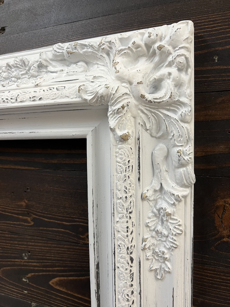 May include: Close-up of a white, ornate picture frame with distressed paint. The frame features detailed floral and scrollwork designs on the corners and edges. The frame is set against a dark wood background.