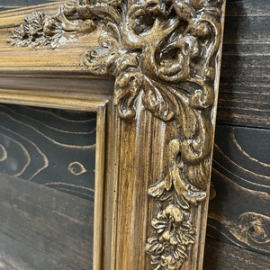 May include: A close-up of a brown ornate picture frame with a detailed, carved design. The frame has a distressed, antique look.