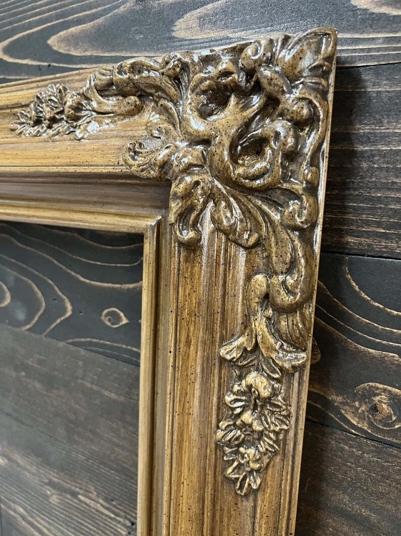 May include: A close-up of a brown ornate picture frame with a detailed, carved design. The frame has a distressed, antique look.