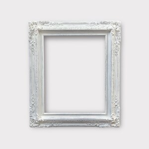 May include: A rectangular, ornate white picture frame with detailed floral and scrollwork carvings. The frame has a distressed, vintage appearance, with visible wear and tear. The frame is empty, ready for a photo or artwork.