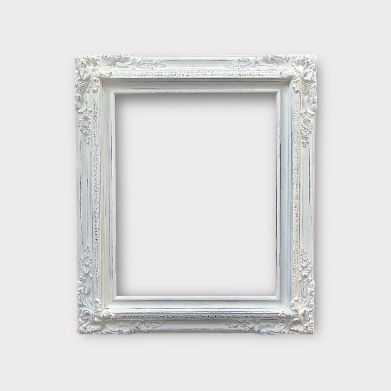 May include: A rectangular, ornate white picture frame with detailed floral and scrollwork carvings. The frame has a distressed, vintage appearance, with visible wear and tear. The frame is empty, ready for a photo or artwork.