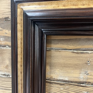 May include: Close-up of a decorative picture frame with a dark brown exterior and a gold-colored inner border. The frame has a detailed, textured design and is set against a wooden background.