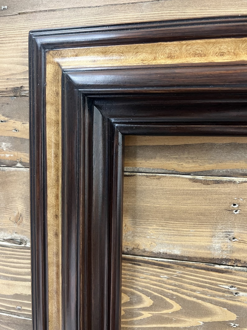 May include: Close-up of a decorative picture frame with a dark brown exterior and a gold-colored inner border. The frame has a detailed, textured design and is set against a wooden background.