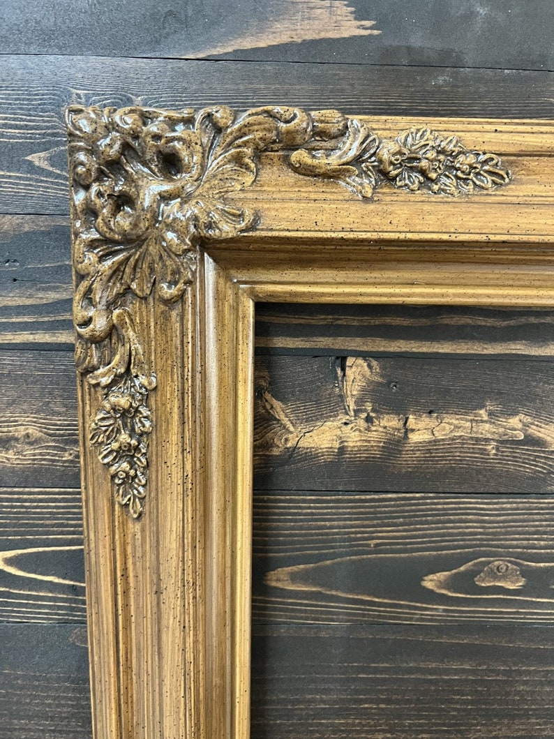May include: A close-up of a brown ornate picture frame with a detailed, carved design. The frame is made of wood and has a distressed finish.