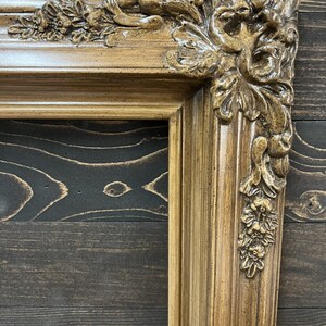 May include: A close-up of a decorative, ornate picture frame with a brown wood finish and intricate carvings. The frame has a distressed look, with a subtle speckled texture.