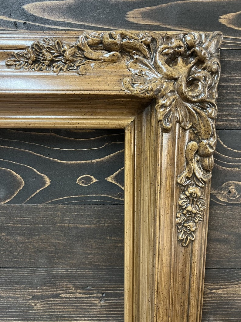 May include: A close-up of a decorative, ornate picture frame with a brown wood finish and intricate carvings. The frame has a distressed look, with a subtle speckled texture.