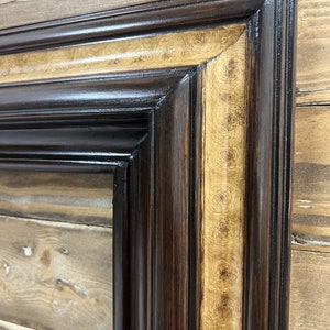 May include: Close-up of a rectangular picture frame with a dark brown exterior and a lighter, patterned interior border. The frame has a glossy finish and is set against a wooden background.