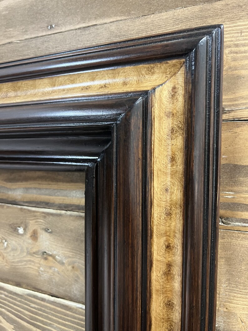 May include: Close-up of a rectangular picture frame with a dark brown exterior and a lighter, patterned interior border. The frame has a glossy finish and is set against a wooden background.