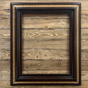 May include: A rectangular picture frame with a dark brown finish and a gold inner border. The frame has a detailed, ornate design and is set against a wooden background.