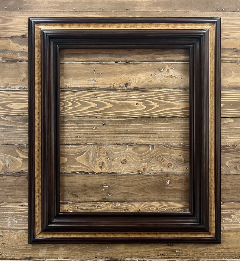 May include: A rectangular picture frame with a dark brown finish and a gold inner border. The frame has a detailed, ornate design and is set against a wooden background.