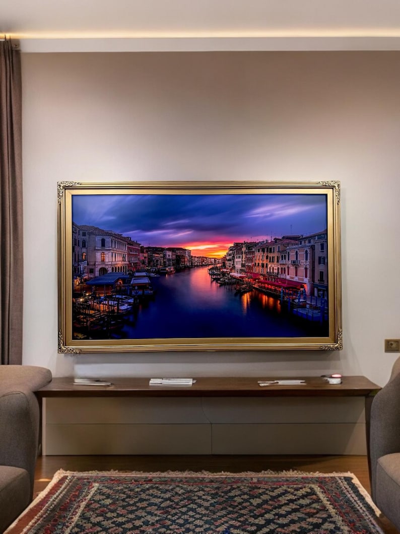 May include: A framed photograph of a cityscape with a canal running through it. The buildings are a mix of yellow, orange, and red, and the water is a deep blue. The sky is a vibrant purple and orange, with clouds in the distance. The frame is a gold color with ornate details.