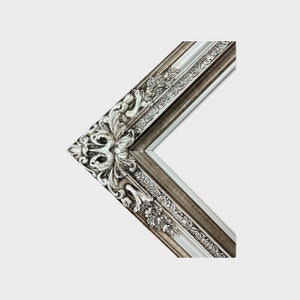 May include: A silver and gray ornate picture frame with a decorative, carved design. The frame has a raised edge and a beveled inner edge.