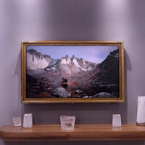 May include: A rectangular landscape photograph of jagged, snow-dusted mountain peaks reflected in a calm lake, encased in an ornate gold frame. The artwork is mounted on a solid gray wall above a smooth, light-brown wooden floating shelf.