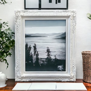 May include: A framed black and white landscape photograph with a white ornate frame. The photograph depicts a misty lake scene with trees in the foreground. The frame is placed on a wooden surface with three white tiles in front.