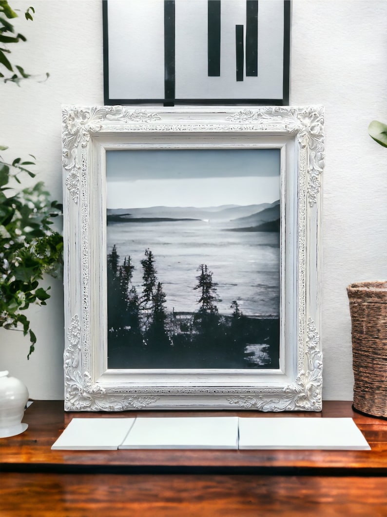 May include: A framed black and white landscape photograph with a white ornate frame. The photograph depicts a misty lake scene with trees in the foreground. The frame is placed on a wooden surface with three white tiles in front.
