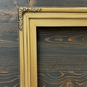 May include: A rectangular picture frame with a metallic gold finish and a smooth, multi-tiered profile. The top left corner features an ornate, raised decorative accent with intricate scrollwork and floral detailing in a darker gold tone.