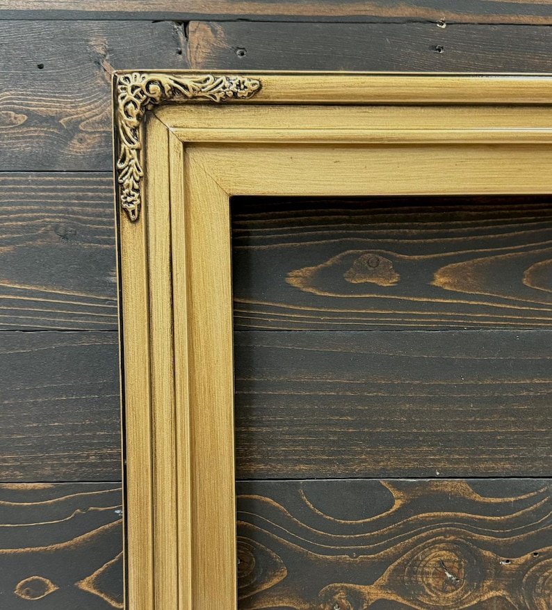 May include: A rectangular picture frame with a metallic gold finish and a smooth, multi-tiered profile. The top left corner features an ornate, raised decorative accent with intricate scrollwork and floral detailing in a darker gold tone.