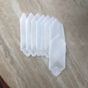 May include: A set of eight white linen napkins with delicate lace trim. The napkins are folded in a triangular shape and arranged in a fan pattern.