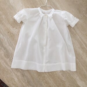 May include: A white cotton baby gown with a ruffled collar and short sleeves. The gown has a button closure down the front.