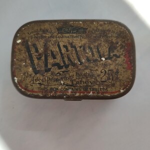 Puede incluir: Caja de caramelos Parola vintage con forma rectangular y tapa con bisagras. La caja es marrón con letras negras y detalles rojos. El texto incluye "Parola" y "Ideal Peppermint Candy". La caja está envejecida y muestra desgaste.