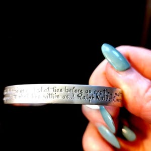 May include: Silver cuff bracelet with an engraved quote by Ralph Waldo Emerson: "What lies before us and what lies within us are tiny matters..." The bracelet is held against a dark background.