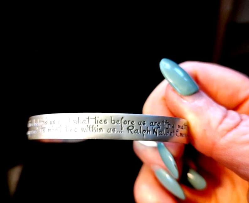 May include: Silver cuff bracelet with an engraved quote by Ralph Waldo Emerson: "What lies before us and what lies within us are tiny matters..." The bracelet is held against a dark background.