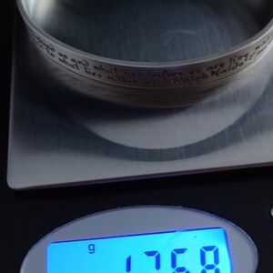 May include: A silver cuff bracelet with an engraved quote by Ralph Waldo Emerson, weighing 17.68 grams. The bracelet is on a digital scale, showing the weight in grams. The text on the bracelet reads: "...what lies before us and what lies within us..."