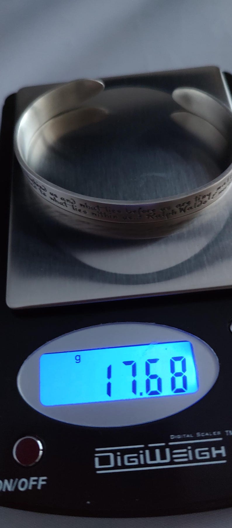 May include: A silver cuff bracelet with an engraved quote by Ralph Waldo Emerson, weighing 17.68 grams. The bracelet is on a digital scale, showing the weight in grams. The text on the bracelet reads: "...what lies before us and what lies within us..."
