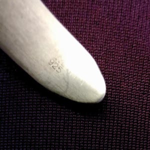May include: Close-up of a polished silver object with the number "925" stamped on its surface. The object has a smooth, rounded shape and is set against a dark purple textured background.