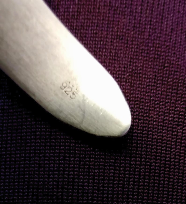 May include: Close-up of a polished silver object with the number "925" stamped on its surface. The object has a smooth, rounded shape and is set against a dark purple textured background.
