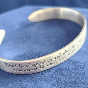 May include: A silver-colored cuff bracelet with an engraved inspirational quote. The text reads "What lies behind us and what lies before us are tiny matters compared to what lies within us." The bracelet is displayed on a blue surface.