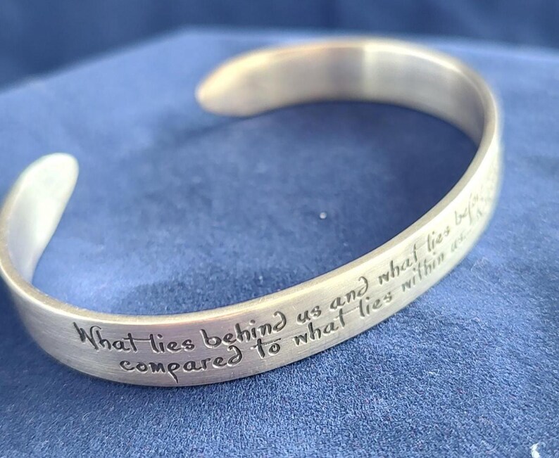 May include: A silver-colored cuff bracelet with an engraved inspirational quote. The text reads "What lies behind us and what lies before us are tiny matters compared to what lies within us." The bracelet is displayed on a blue surface.