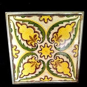 Op de afbeelding: Vierkante keramische tegel met een bloemmotief in geel, bruin en groen, gezet in een zilveren metalen frame. De tegel heeft een symmetrisch patroon van gestileerde bladeren en bloemen, ideaal voor interieurdecoratie.