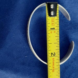 May include: A silver cuff bracelet with a textured design, measured with a yellow tape measure. The bracelet is approximately 1.75 inches in diameter. The background is a solid blue fabric.
