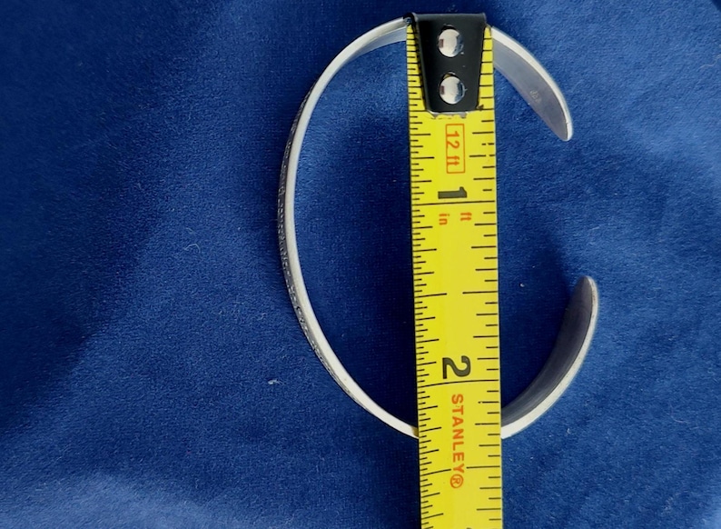 May include: A silver cuff bracelet with a textured design, measured with a yellow tape measure. The bracelet is approximately 1.75 inches in diameter. The background is a solid blue fabric.