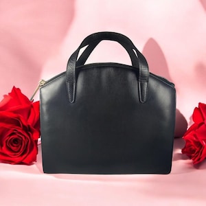 May include: A black leather handbag with a top zipper closure and two short handles. The bag has a structured shape and a gold-colored zipper. Two red roses are on either side of the bag against a pink background.