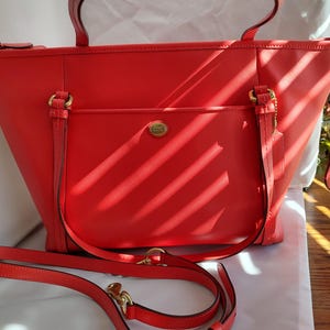 May include: A bright red leather tote bag with a matching shoulder strap. The bag features a front pocket, gold-tone hardware, and top handles. The Coach logo is visible on the front.
