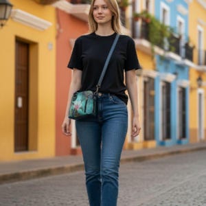 Puede incluir: Una persona con una camiseta negra y vaqueros azules camina por una calle adoquinada. Lleva un bolso cruzado turquesa con un diseño floral. El fondo presenta edificios coloridos.