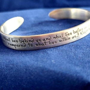 May include: A silver-colored cuff bracelet with engraved text. The inscription reads, "What lies behind us and what lies before us compared to what lies within us..." The bracelet is on a blue surface.